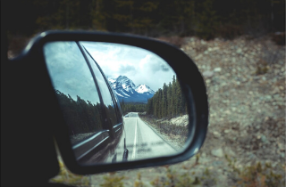 A car side mirror looks back on a long road, surrounded by forest, reflecting on change.