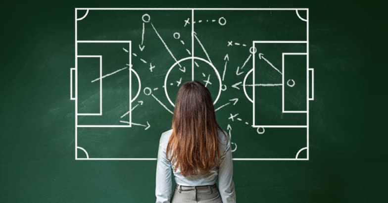 A female Bid Consultant looks at a soccer strategy on a chalkboard, comparing it to a bidding strategy